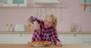 Pembe tişörtlü şirin sarışın kız pembe mutfakta elleriyle pizza yiyor. Aç liseli kız erimiş peynirle pizza yemekten hoşlanır..