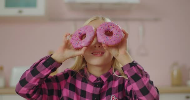 Söt blond liten flicka i rosa skjorta med två rosa munkar i händerna i ansiktet. Rosa munkar ögon. Roligt koncept med sötsaker. — Stockvideo