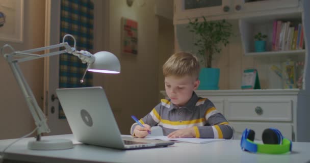 Vorschulkind lernt online zu Hause. Online-Bildung und Fernunterricht für Kinder. Junge hört Lehrauftrag vom Laptop aus und schreibt in Notizbuch. — Stockvideo