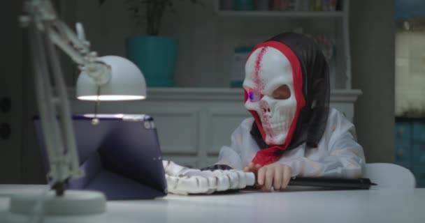 Barn i halloween skalle kostym med blinkande ögon skrämmande vänner med skrämmande hand genom videosamtal på surfplatta dator. Karantänhalloween firande. — Stockvideo