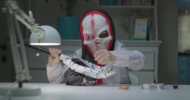 Kid in halloween skull costume with flashing eye examining how many sweets he collected. Trick or treat. Lot of candies from halloween bag. — Stock Video