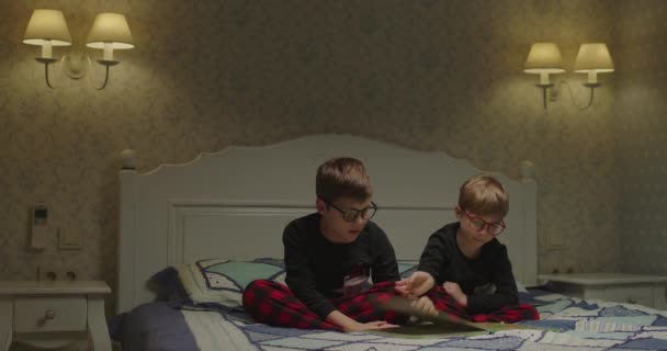 Dvě děti v brýlích a stejné pyžamo na čtení knihy před spaním. Bratři otáčejí stránky papírové knihy s obrázky. Dítě s problémy se zrakem. — Stock video