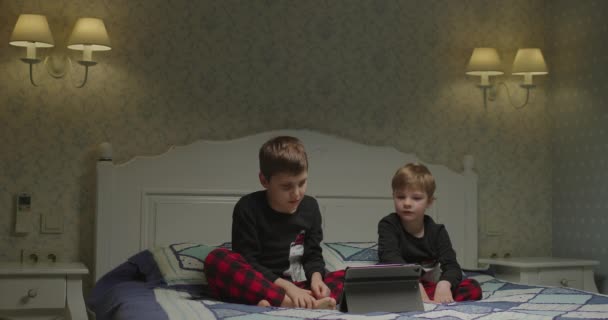 Due bambini che usano tablet sul letto nella camera dei genitori la sera. Fratelli a guardare gadget insieme. — Video Stock