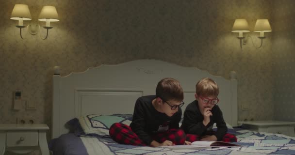 Broers en zussen lezen papieren boek met foto 's thuis' s avonds zittend op bed. Kinderen lezen graag samen het boek. Broeders in dezelfde kleren. — Stockvideo