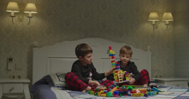 Dos chicos en el mismo pijama jugando con bloques de construcción de plástico de color sentados en la cama por la noche. Niños jugando antes de irse a la cama. Los hermanos juegan juntos en casa. — Vídeos de Stock