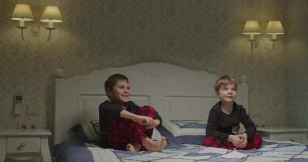 Twee broers in dezelfde pyjama wachtend op de val van plastic bouwstenen. Speelgoed valt op het bed. broers en zussen die samen spelen. — Stockvideo