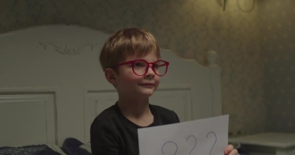 Söt elementär ålder unge i glasögon hålla ark av papper med frågetecken skrivna på den. Att hitta en lösning på problemet. — Stockvideo