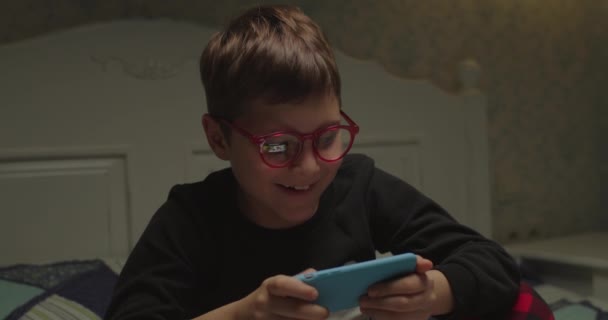 Niño de la escuela en gafas de los ojos jugando videojuegos en el teléfono inteligente. Chico usando artilugio a la hora de acostarse. Niño disfruta viendo dibujos animados en línea en el teléfono móvil. — Vídeo de stock