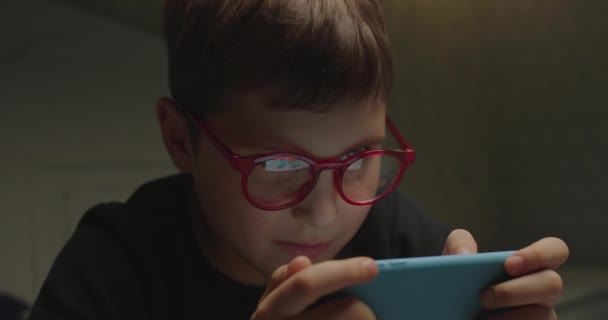 Close up of schoolboy in eye glasses playing video games on smartphone. Kluk, co používá gadget před spaním. Dítě si užívá sledování karikatur online na mobilním telefonu. — Stock video