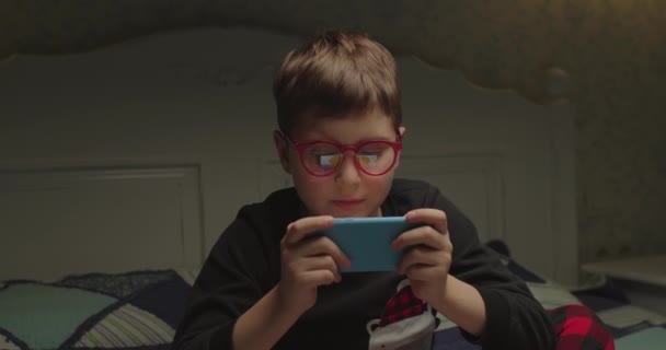 Ένα έφηβο παιδί με πιτζάμες και γυαλιά να βλέπει εφαρμογή κινητού στο κρεβάτι. Αγόρι που παίζει παιχνίδια στο σπίτι. — Αρχείο Βίντεο