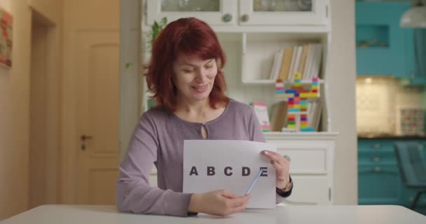 Nauczycielka po 40-tce rozmawia z kamerą i trzyma kartkę papieru z literami alfabetu. Edukacja online i proces uczenia się na odległość. Praca w domu nauczyciel angielskiego. — Wideo stockowe