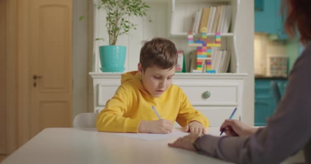 Rapaz da escola a fazer trabalhos de casa com o terapeuta. Criança com autismo escrevendo com caneta com professor ajudando-o. Processo de educação em autismo. Ensino em casa. — Vídeo de Stock