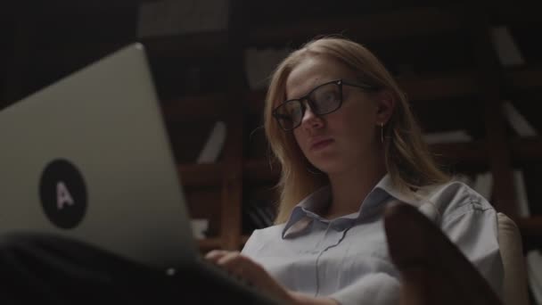 暗い図書館のアームチェアに座ってノートパソコンで働く20代の女性。女性のフリーランサーがノートパソコンに入力し、エッセイを書く。家庭での千年紀の勉強。オンライン作業と教育の概念. — ストック動画