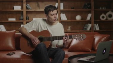 20 'li yaşlarda bir adam evdeki kanepede oturup dizüstü bilgisayara bakarak gitar çalmayı öğreniyor. Çevrimiçi müzik öğrenme kavramı. Müzik öğrencisi akustik gitar çalar.