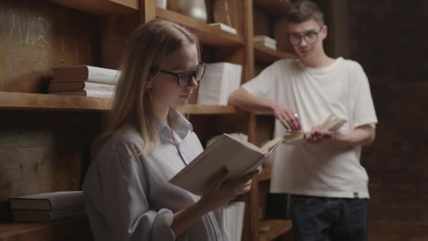 Joven pareja inteligente en anteojos leyendo libros en la biblioteca. Los estudiantes de 20 disfrutan leyendo libros de papel juntos. Elegante estantería de madera. — Vídeos de Stock