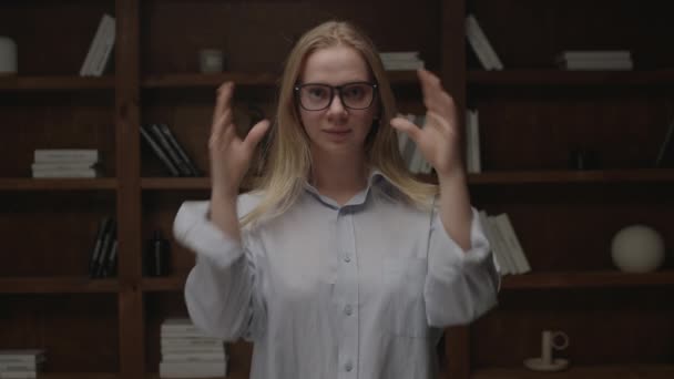 Portrait of young blonde woman taking off and on eye glasses, smiling and looking at camera standing in dark room. Millennial student enjoys studying. — Stock Video