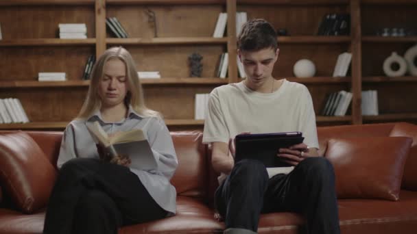 Giovane moglie che legge libro cartaceo e marito che legge libro elettronico su computer tablet. 20s coppia lettura seduta sul divano a casa. — Video Stock