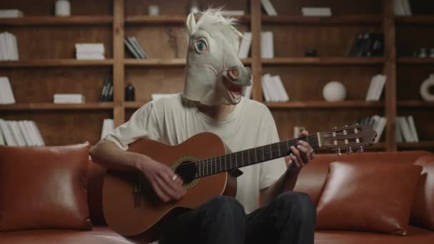 Persona con testa di unicorno che suona la chitarra. Musica buffa. Unicorno suona la chitarra seduta sul divano. — Video Stock