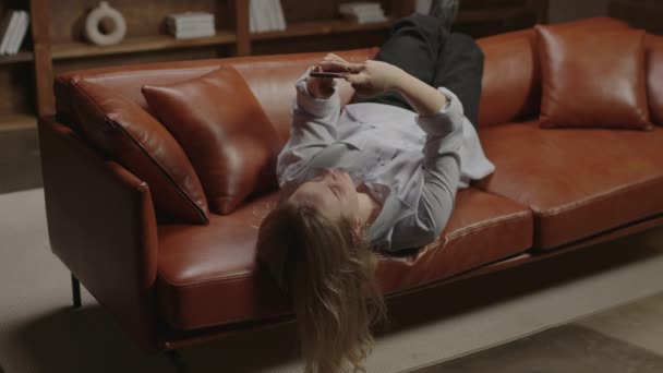 20代の女性がネットサーフィンや携帯電話でのテキストメッセージをソファに寝そべって。ソファに寝そべっている若い女性。ガジェット中毒千年. — ストック動画