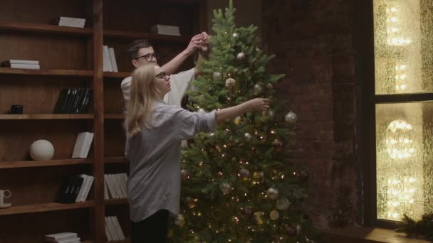 A 20-as évekbeli fiatalok karácsonyfát díszítenek együtt az otthoni könyvtárban. Szerető férfi és nő szemüvegben ölelés közel karácsonyfa. — Stock videók