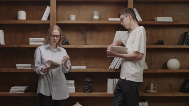 20-talsstudenter läser böcker på biblioteket. Unga smarta par tycker om att läsa pappersböcker tillsammans. Snygg bokhylla i trä. — Stockvideo