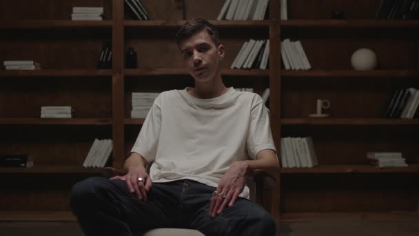 20 anni uomo sicuro di sé in t-shirt bianca seduto in poltrona e biblioteca e guardando la fotocamera a tarda notte. — Video Stock