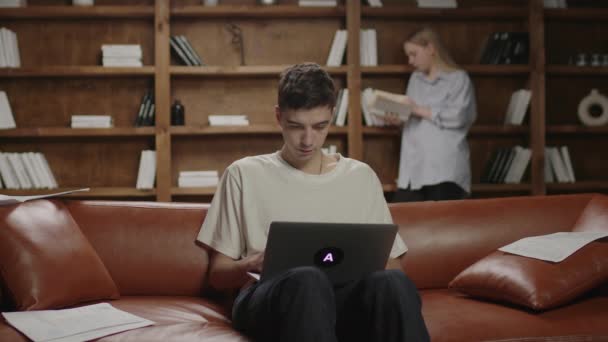 Estudiante masculino escribiendo en el ordenador portátil, mujer está leyendo libro detrás. Un par de estudiantes en casa. Gente inteligente escribiendo y leyendo. — Vídeo de stock