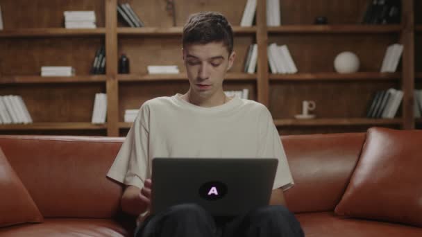 20多岁的年轻人坐在沙发上在笔记本电脑上打字。男性自由职业者关闭笔记本电脑和思考。千年在家里学习。在线工作和教育. — 图库视频影像