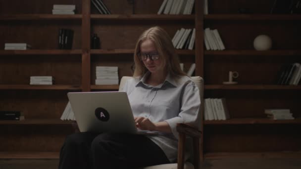 Mujer joven de 20 años trabajando en un portátil sentado en un sillón en la biblioteca oscura. Freelancer femenina escribiendo en el portátil, escribiendo ensayo. Millennial estudiando en casa. Concepto de trabajo y educación en línea. — Vídeos de Stock
