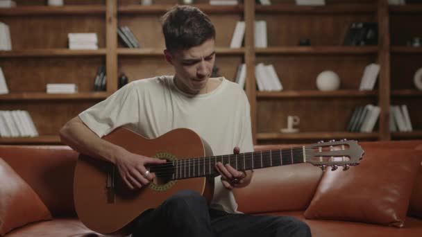 Hombre joven de 20 años tocando la guitarra sentado en el sofá en casa. Profesor de guitarra hablando con los estudiantes mirando la cámara. Concepto de aprendizaje de música online. — Vídeo de stock