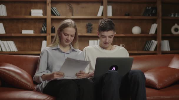 Dwóch tysiącletnich studentów piszących esej, kobieta dyktująca informacje na kartce papieru, mężczyzna piszący na laptopie. Praca zespołowa studentów na kanapie. — Wideo stockowe