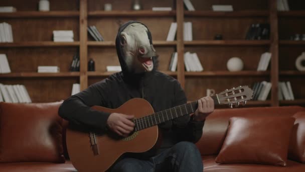 Person in lustiger Maske des weißen Pferdes, die auf dem Sofa sitzend Gitarre spielt. Einhorn mit Gitarre. Lustige Musik. — Stockvideo