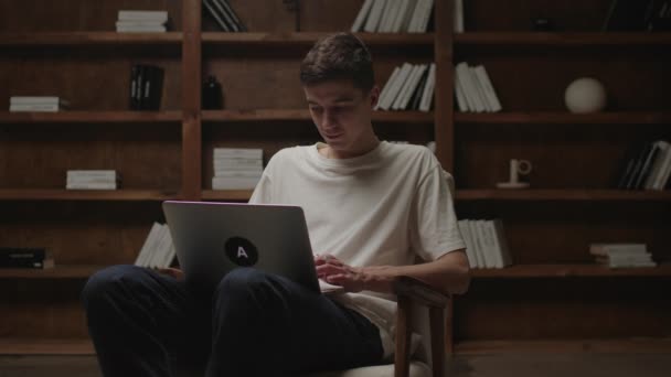 Молоді 20-ти чоловік на ноутбуці сидять у кріслі. Тисячолітня школа в домашній бібліотеці. Онлайн-робота та концепція освіти. — стокове відео