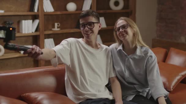 Doi tineri bloggeri mileniali care înregistrează videoclipuri pe camera de filmat stând pe canapea. Bloggeri inteligenți cuplu în ochelari de ochi vorbind cu abonații lor . — Videoclip de stoc