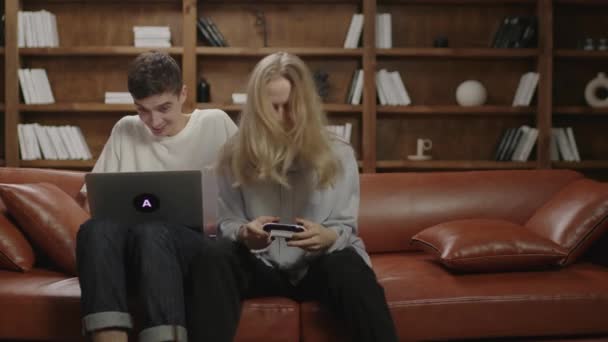 Millennial kvinna spelar tv-spel medan hennes pojkvän arbetar på laptop hemifrån. Ung familj som tillbringar kvällstid tillsammans på soffan. Fru stör förvärvsarbetande make. — Stockvideo