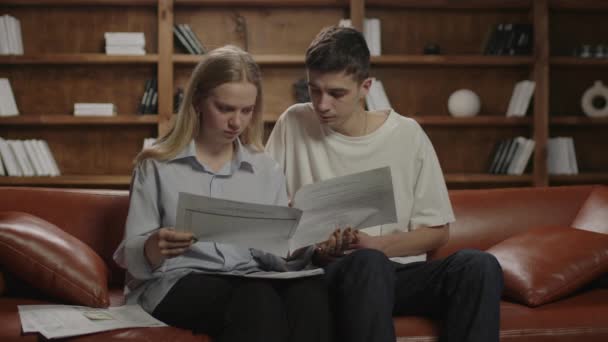 Pareja joven revisando facturas de papel y documentos domésticos, calculando gastos y planificando el presupuesto familiar sentada en el sofá. — Vídeos de Stock