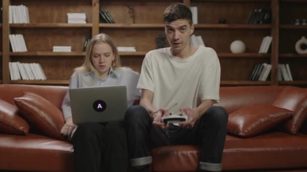Kobieta pracująca na laptopie z chłopakiem przeszkadza jej grając w gry wideo. Młoda rodzina spędza razem wieczór na kanapie. Żona pracuje, mąż gra.. — Wideo stockowe