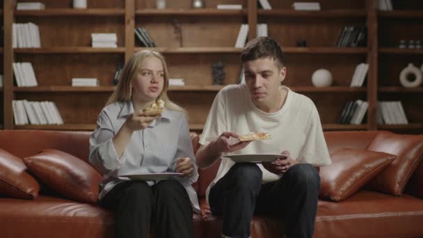 Millennial couple eating pizza and watching TV sitting on couch at home. Young family eating delivery food. — Stock Video