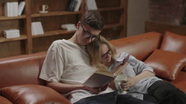 Mąż czyta książki papierowe i żona za pomocą telefonu komórkowego surfing internet. Piękny millenium rodzina przytulanie spędzać wieczór razem na kanapie w domu. — Wideo stockowe