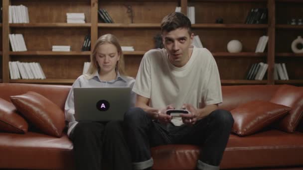 Tysiącletni mężczyzna gra w gry wideo, podczas gdy jego dziewczyna pracuje na laptopie w domu. Młoda rodzina spędza razem wieczór na kanapie. Żona pracuje, mąż gra.. — Wideo stockowe