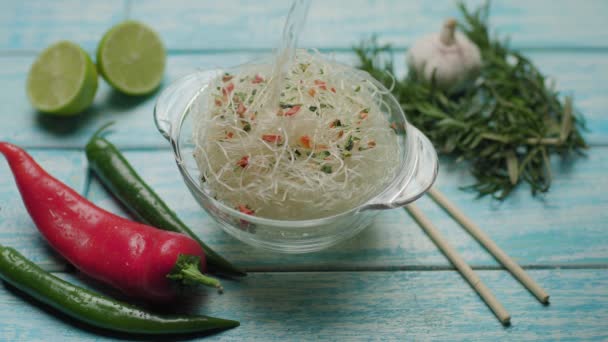 Hot boiling water pouring into glass bowl with asian noodles with vegetables. Instant food cooking. Top view of noodle soup and spices on the blue table. — Stock Video