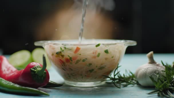 Acqua calda versando in una ciotola di vetro con tagliatelle asiatiche con verdure. Cottura istantanea dei noodle. Vista laterale della ciotola di vetro con zuppa di tagliatelle e spezie sul tavolo blu. — Video Stock