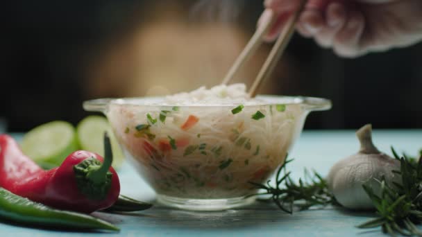 Mano con palillos mezclando fideos de arroz en un tazón de vidrio. Cocina instantánea de fideos. Sopa de fideos y especias en la mesa azul. Plato caliente al vapor. — Vídeo de stock