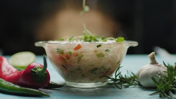 Cadendo micro verdi in ciotola di vetro con tagliatelle asiatiche con verdure. Cottura istantanea dei noodle. Vista laterale della ciotola di vetro con zuppa di tagliatelle e spezie sul tavolo blu. — Video Stock