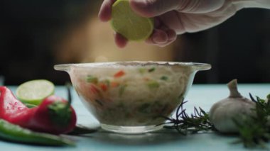 Sebzeli Asya eriştesiyle cam kaseye limon suyu sıkıyorum. Anında şehriye pişirme. Mavi masanın üzerinde erişte çorbası ve baharat olan cam kasenin yan görüntüsü..