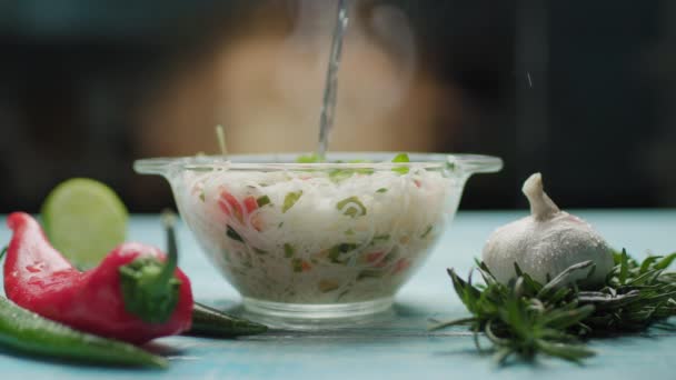Água quente derramando em tigela de vidro com macarrão asiático com legumes. Cozinhar macarrão instantâneo. Vista lateral da tigela de vidro com sopa de macarrão e especiarias na mesa azul. — Vídeo de Stock