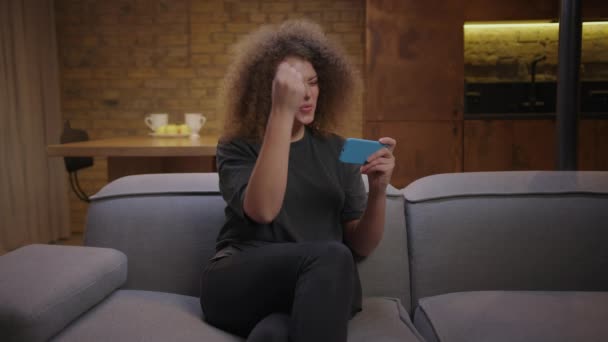 Millennial vrouw spelen mobiele games op mobiele telefoon en het winnen van het spel zittend op de bank. Vrouw maken overwinning gebaar met de handen tijdens het spelen op mobiele telefoon. — Stockvideo