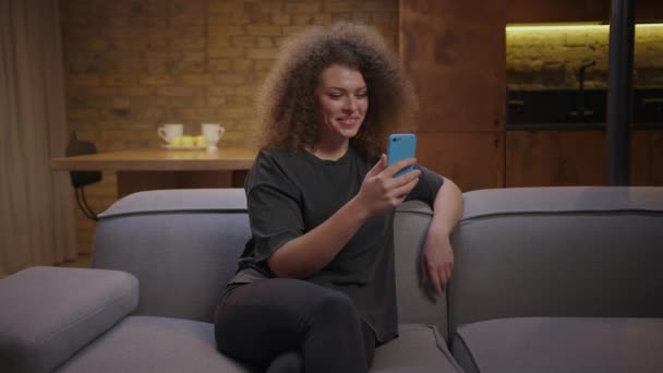 Kobieta z kręconymi włosami, rozmawiająca przez wideo połączenie trzymając komórkę w ręku. Młoda dorosła kobieta korzystająca z telefonu komórkowego do komunikacji online. Tysiąclecie z gadżetem. — Wideo stockowe