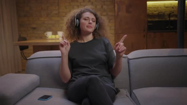 Mujer joven de 20 años disfruta escuchando música en auriculares inalámbricos con los ojos cerrados sentados en el sofá en casa. Mujer guapa rizada con auriculares escuchando la música y bailando con las manos. — Vídeo de stock