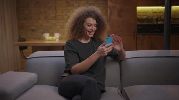 Mladá žena mluví videohovory pomocí mobilního telefonu, který sedí doma na gauči. 20s žena chatování s přítelem on-line. — Stock video